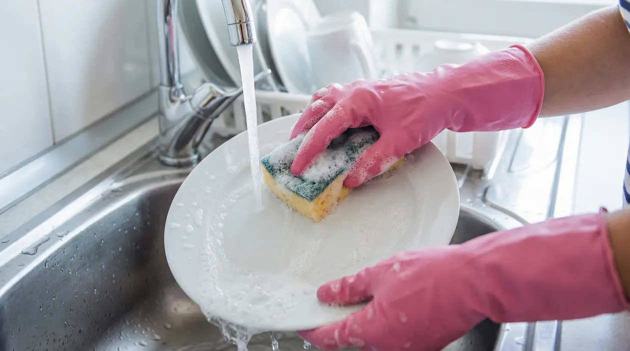 Persona con guanti rosa lava un piatto insaponato sotto l'acqua corrente