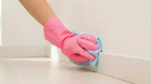 Mano con guanto rosa che pulisce un battiscopa bianco con un panno azzurro