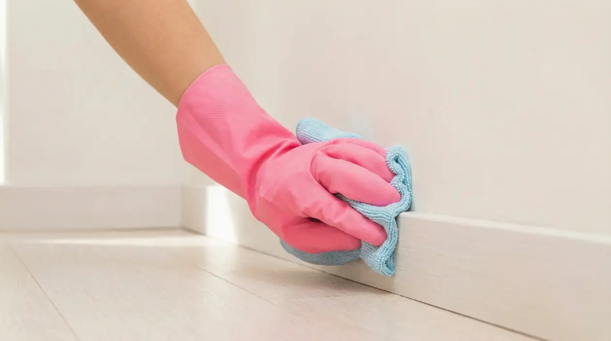 Mano con guanto rosa che pulisce un battiscopa bianco con un panno azzurro